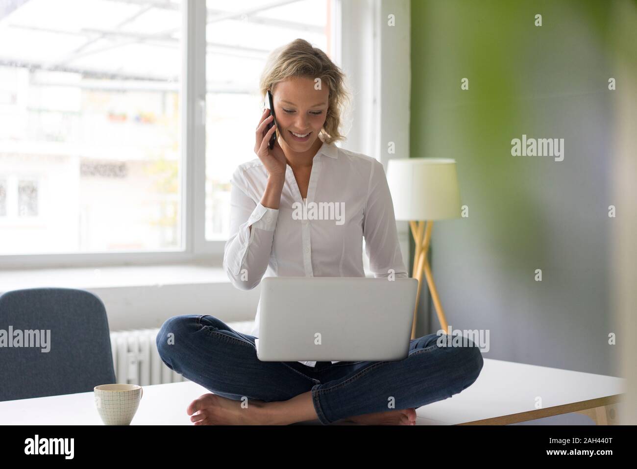 Sorridente giovane imprenditrice seduta sulla scrivania in ufficio utilizzando computer portatile e un telefono cellulare Foto Stock