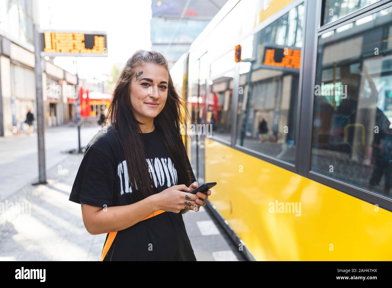 Ritratto di tatuati giovane donna con lo smartphone in piedi alla fermata del tram, Berlino, Germania Foto Stock
