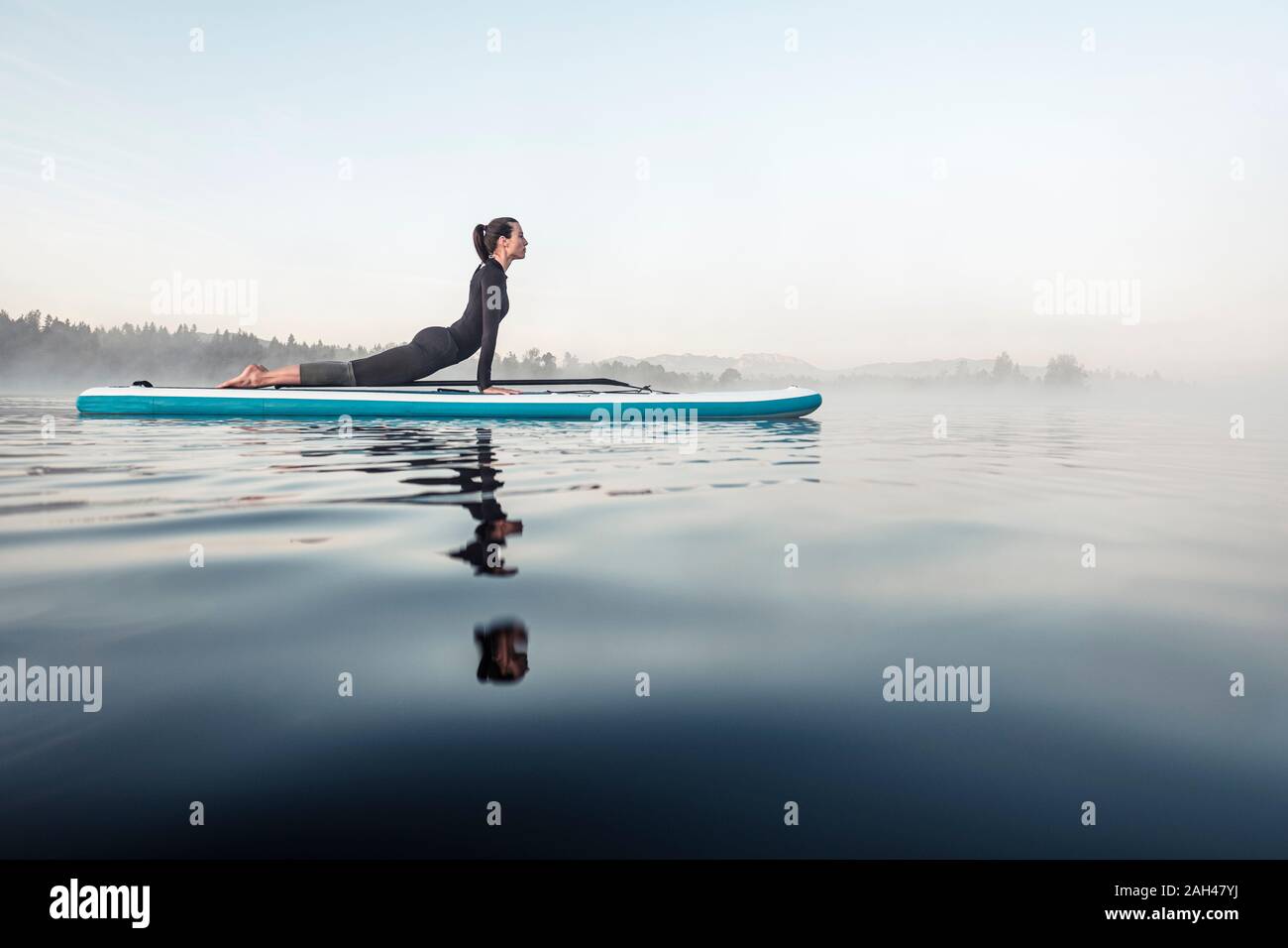 Donna pratica di paddle board yoga sul lago Kirchsee al mattino, Bad Toelz, Baviera, Germania Foto Stock