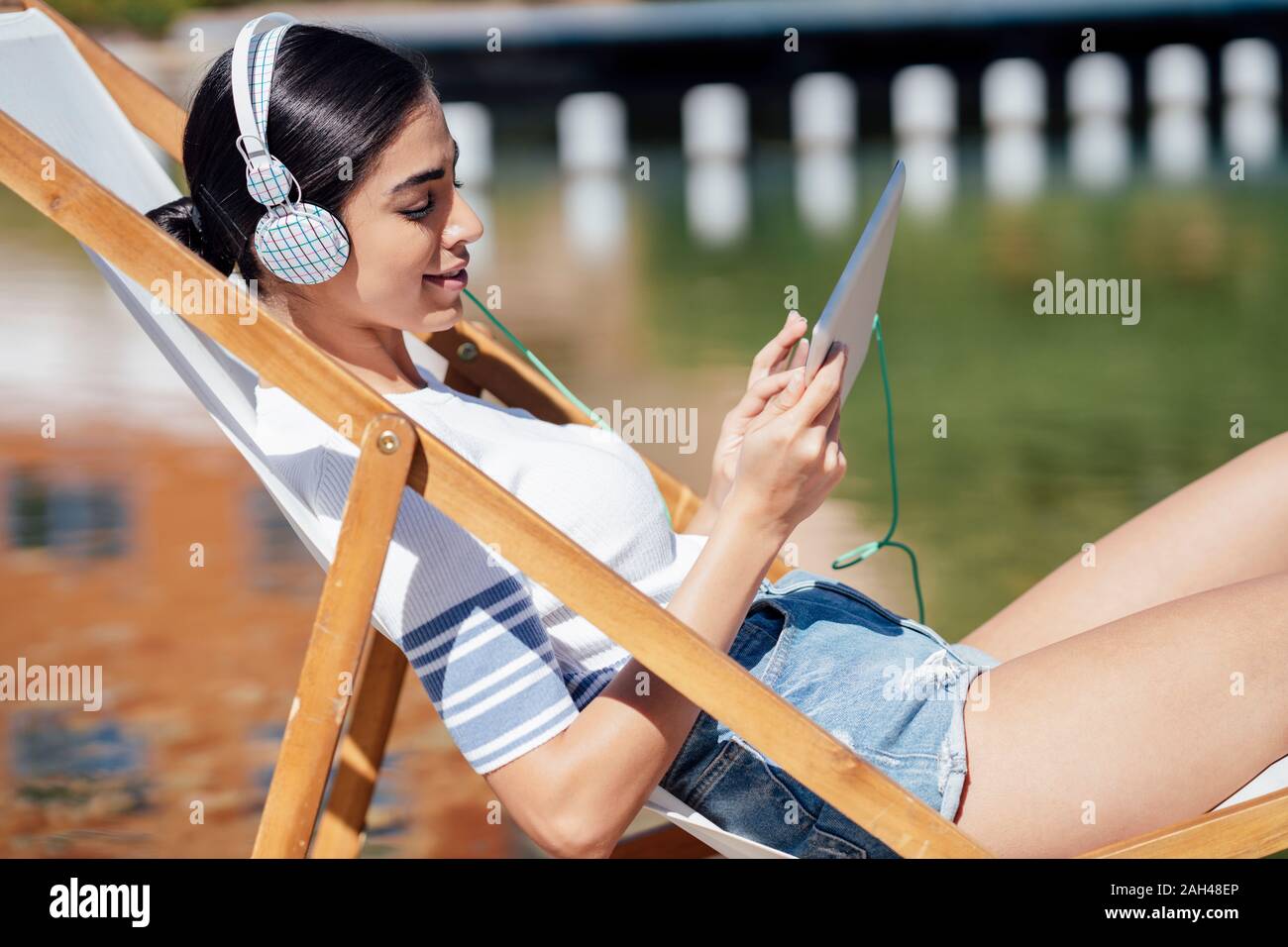 Giovane donna seduto in una sedia a sdraio che indossa le cuffie e utilizzando tablet Foto Stock