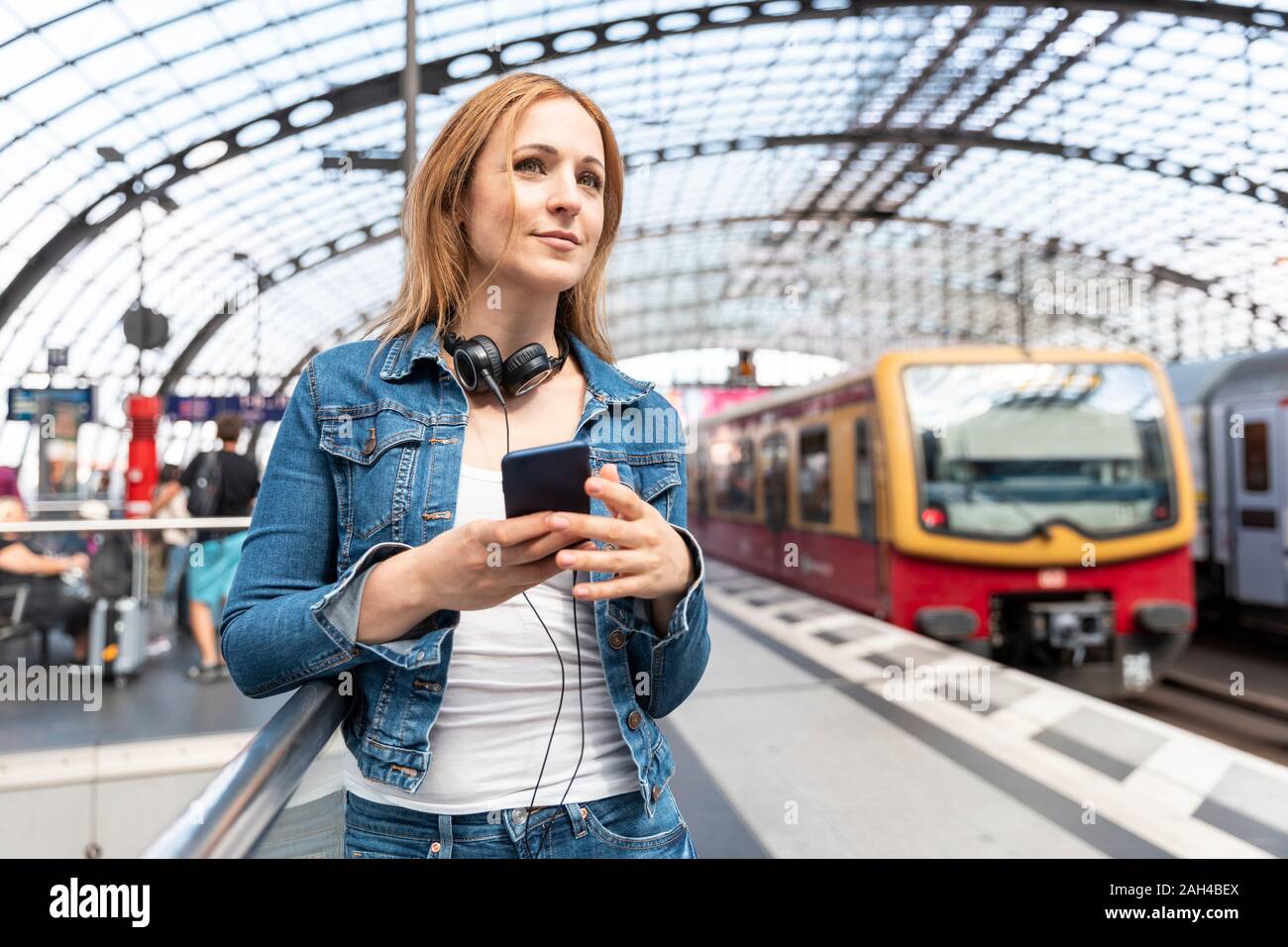 Donna sorridente con lo smartphone e le cuffie sulla piattaforma della stazione, Berlino, Germania Foto Stock