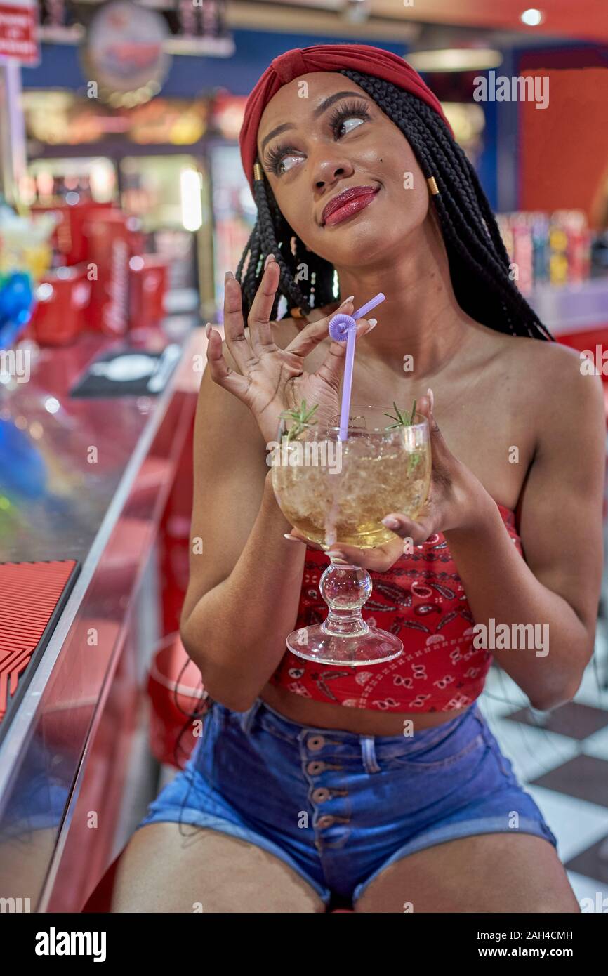 Giovane donna con capelli intrecciati seduto su una barra, bere un cocktail con una paglia Foto Stock