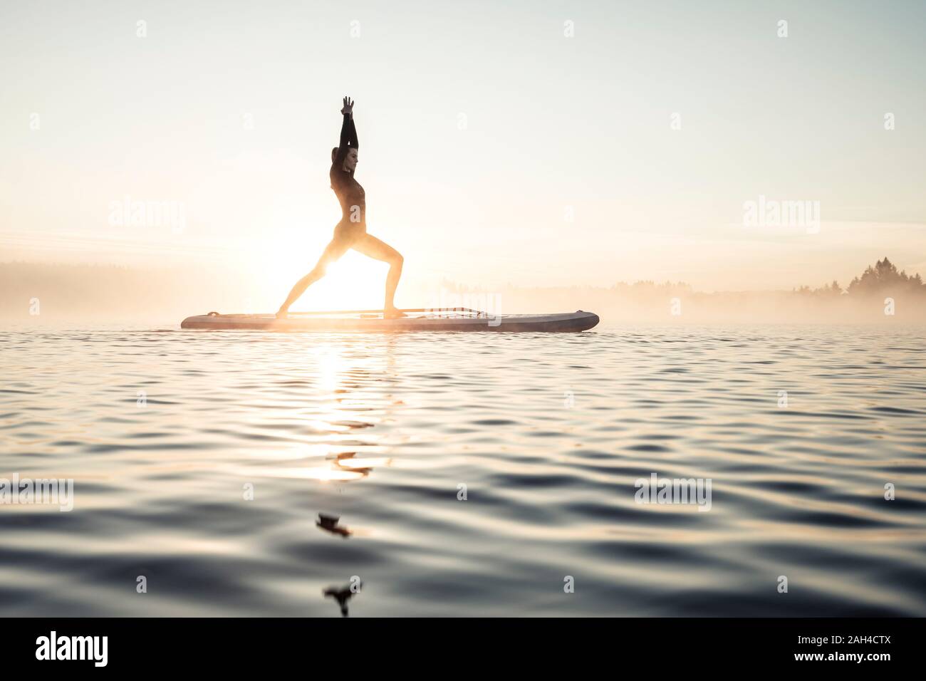 Donna pratica di paddle board yoga sul lago Kirchsee al mattino, Bad Toelz, Baviera, Germania Foto Stock
