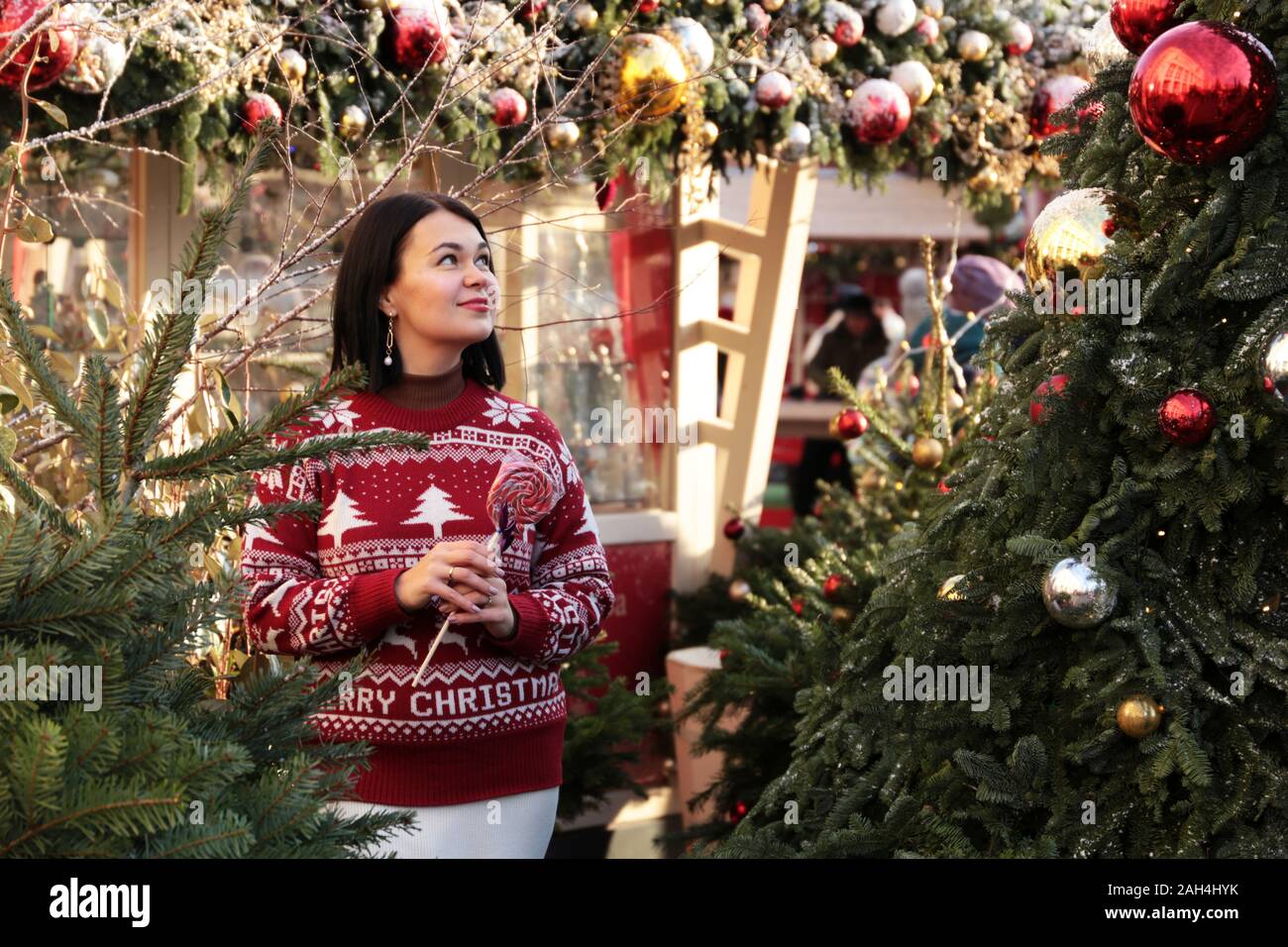 Donna felice in un maglione rosso con lollipop gode la magia delle vacanze di Natale in piedi su una strada sul nuovo anno sfondo ad albero Foto Stock
