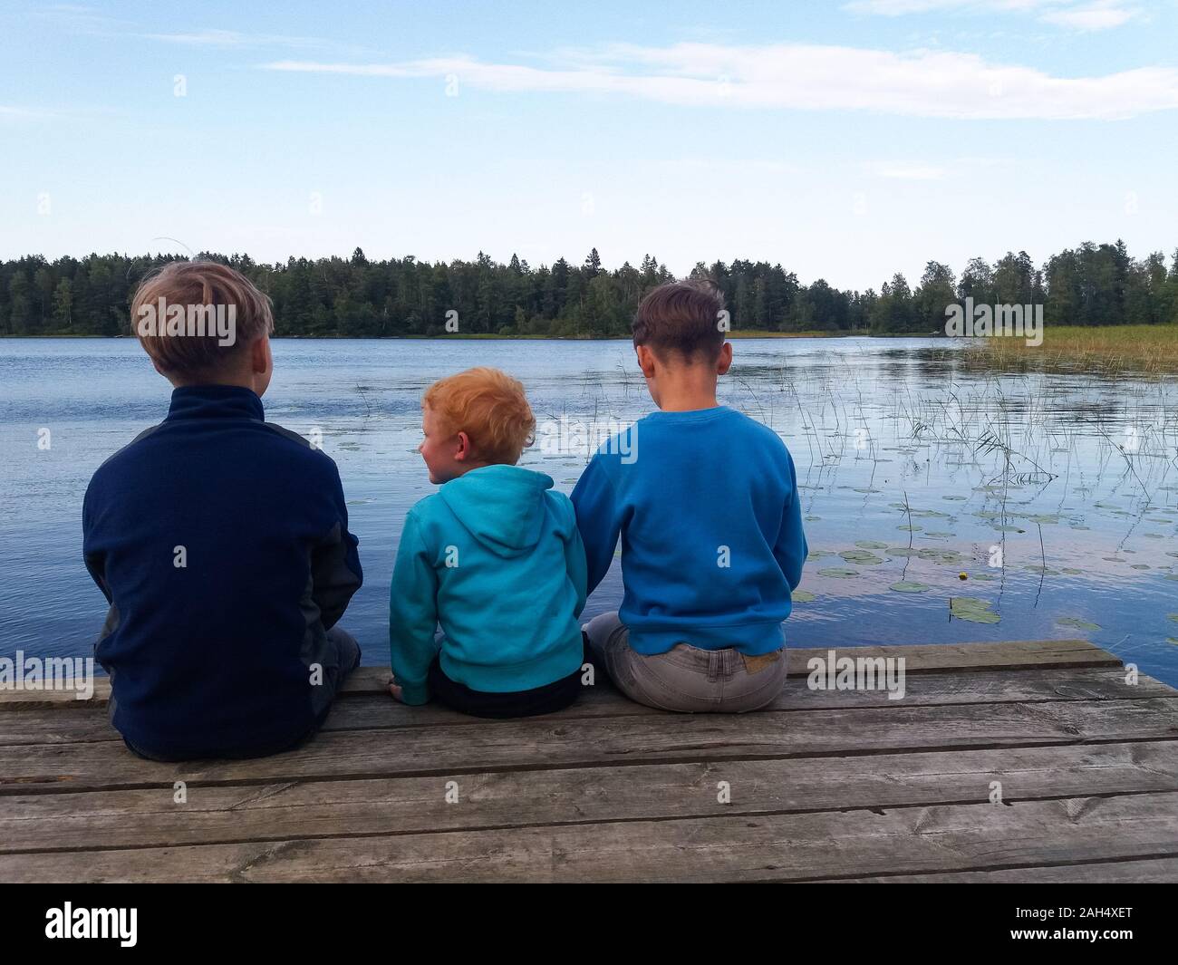 Tre ragazzi di diversa età in blu maglioni di diversi colori sono seduti su di un legno caldo pontone, gambe penzolanti, le spalle al visualizzatore, guardando il lago e la foresta verde dietro di esso. Foto Stock