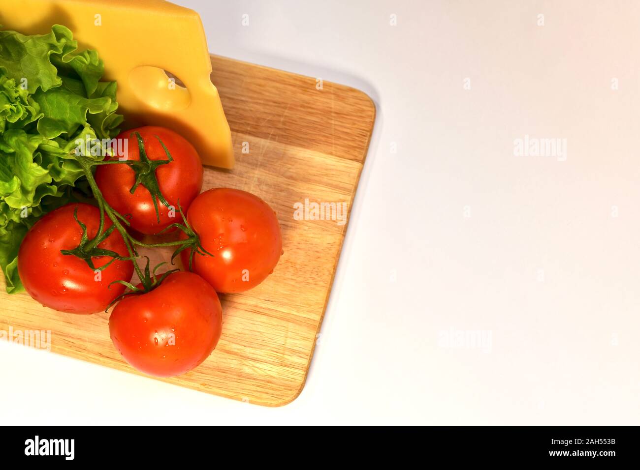 Un insieme di prodotti freschi nelle vicinanze, formaggio, pomodori e lattuga verde su un tagliere di legno su uno sfondo luminoso. Foto Stock