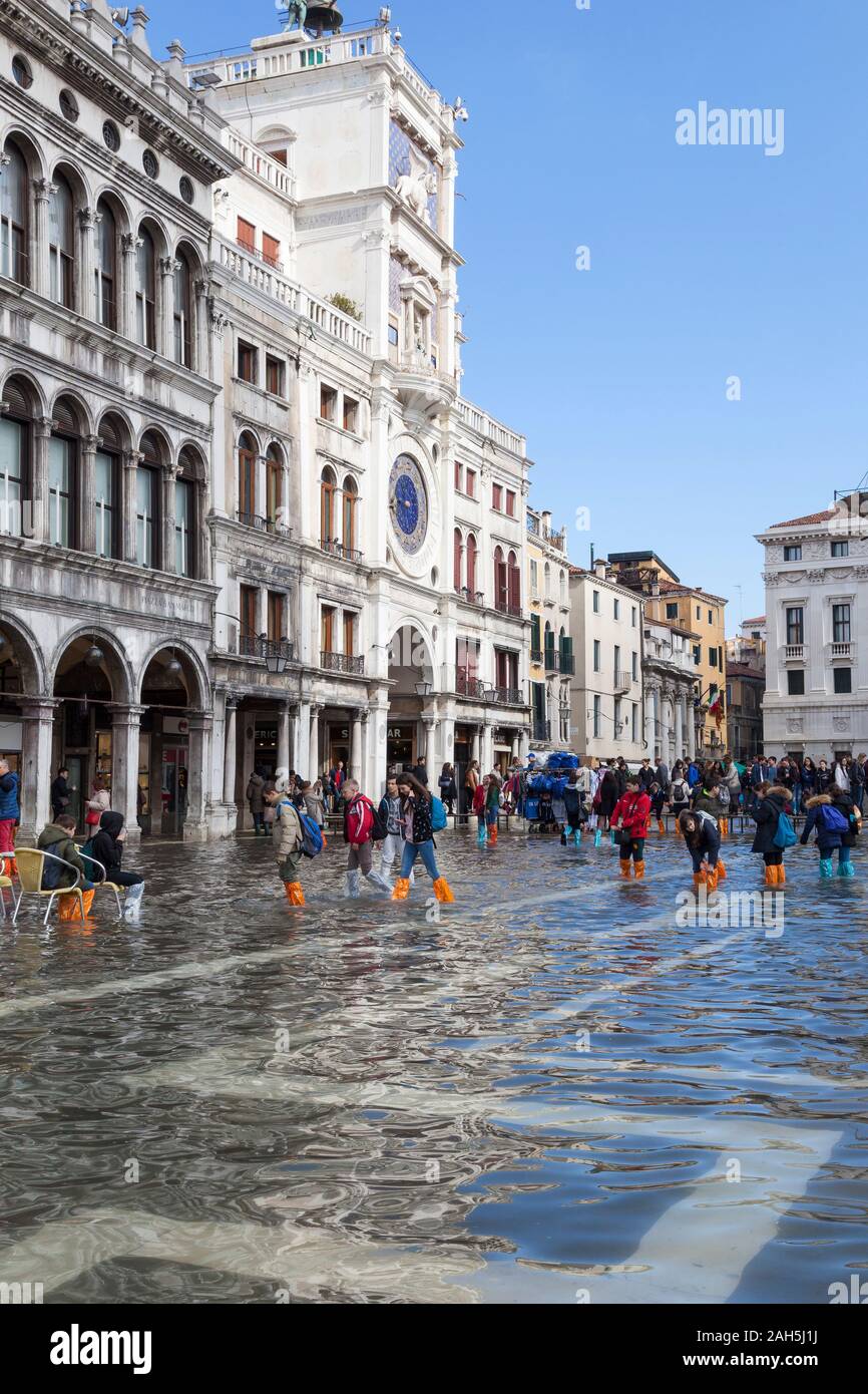 Acqua Alta inondazioni durante l estrema alta marea Piazza San Marco, Venezia, Italia con i turisti in colorate stivali guadare in acqua di inondazione nei pressi di Torre dell Orologio Foto Stock