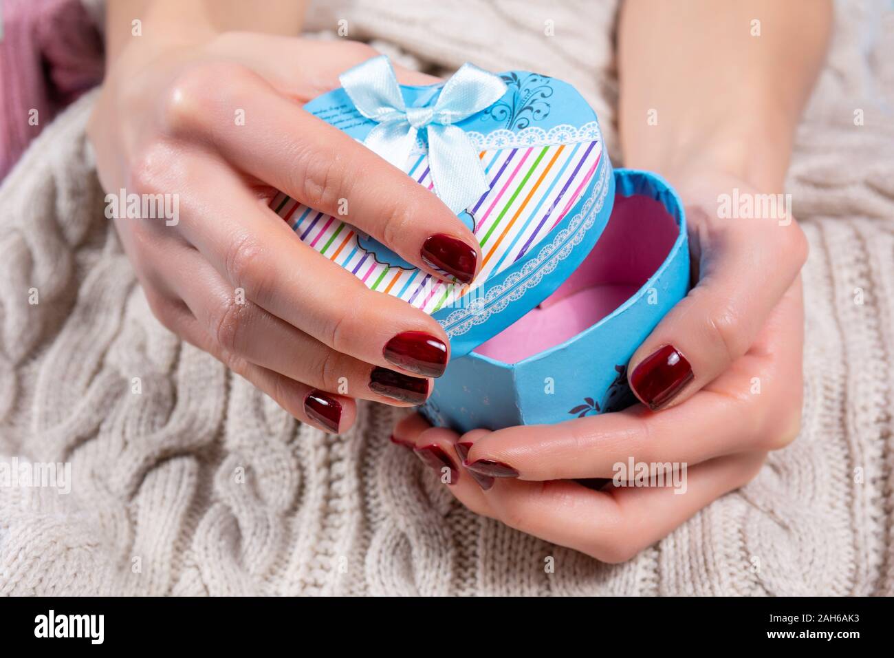 Ragazza mani e apertura di blue box regalo in forma di cuore con un tirante sulla prua maglione di lana sfondo. Mani femminili con chiodi marroni polacco Foto Stock