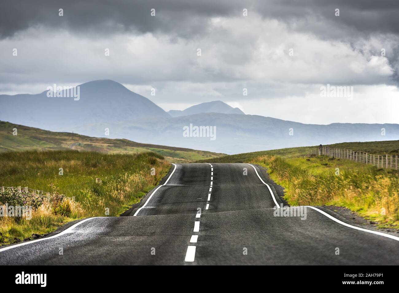 Vista simmetrica di una stretta e ondulata strada di campagna scozzese che si estende fino all'orizzonte, sotto un cielo scuro nuvoloso Foto Stock