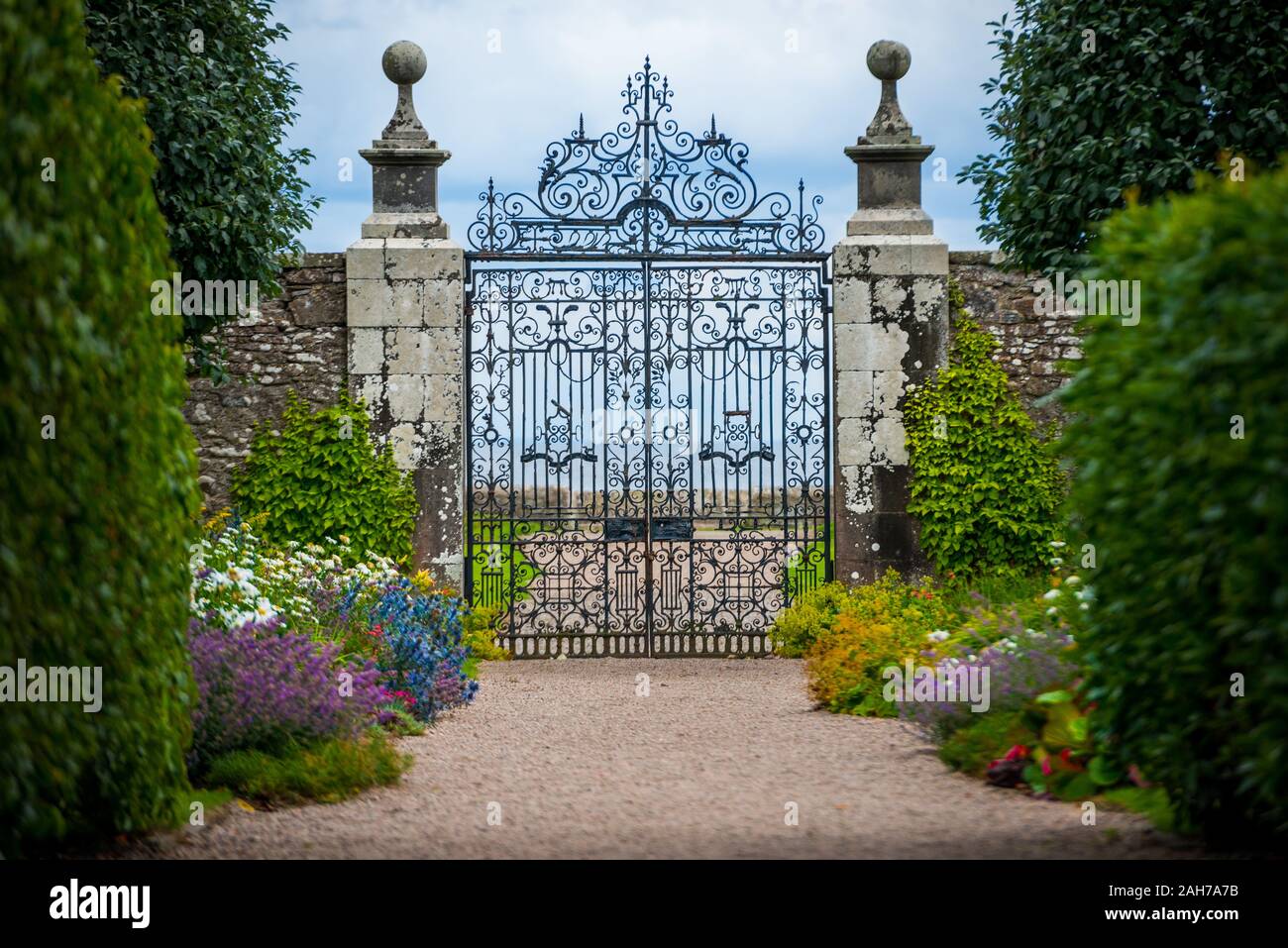 Primo piano di un antico cancello in ferro battuto che conduce un vicolo fiancheggiato da colorati cespugli di fiori e verde siepi Foto Stock