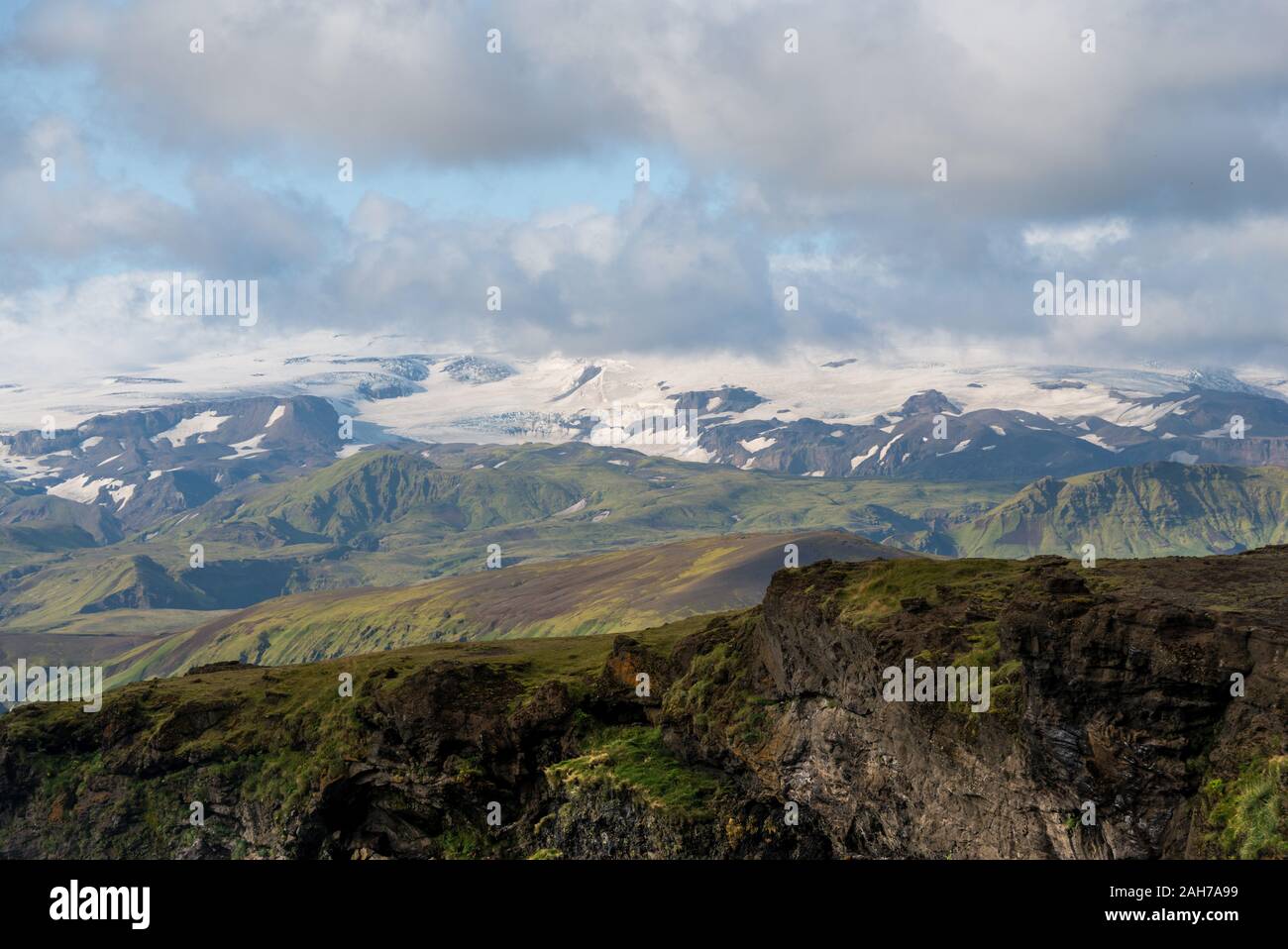 Paesaggio islandese con una dorsale di montagna verde in primo piano, e distanti montagne innevate in lontananza, sotto un cielo blu con le nuvole Foto Stock