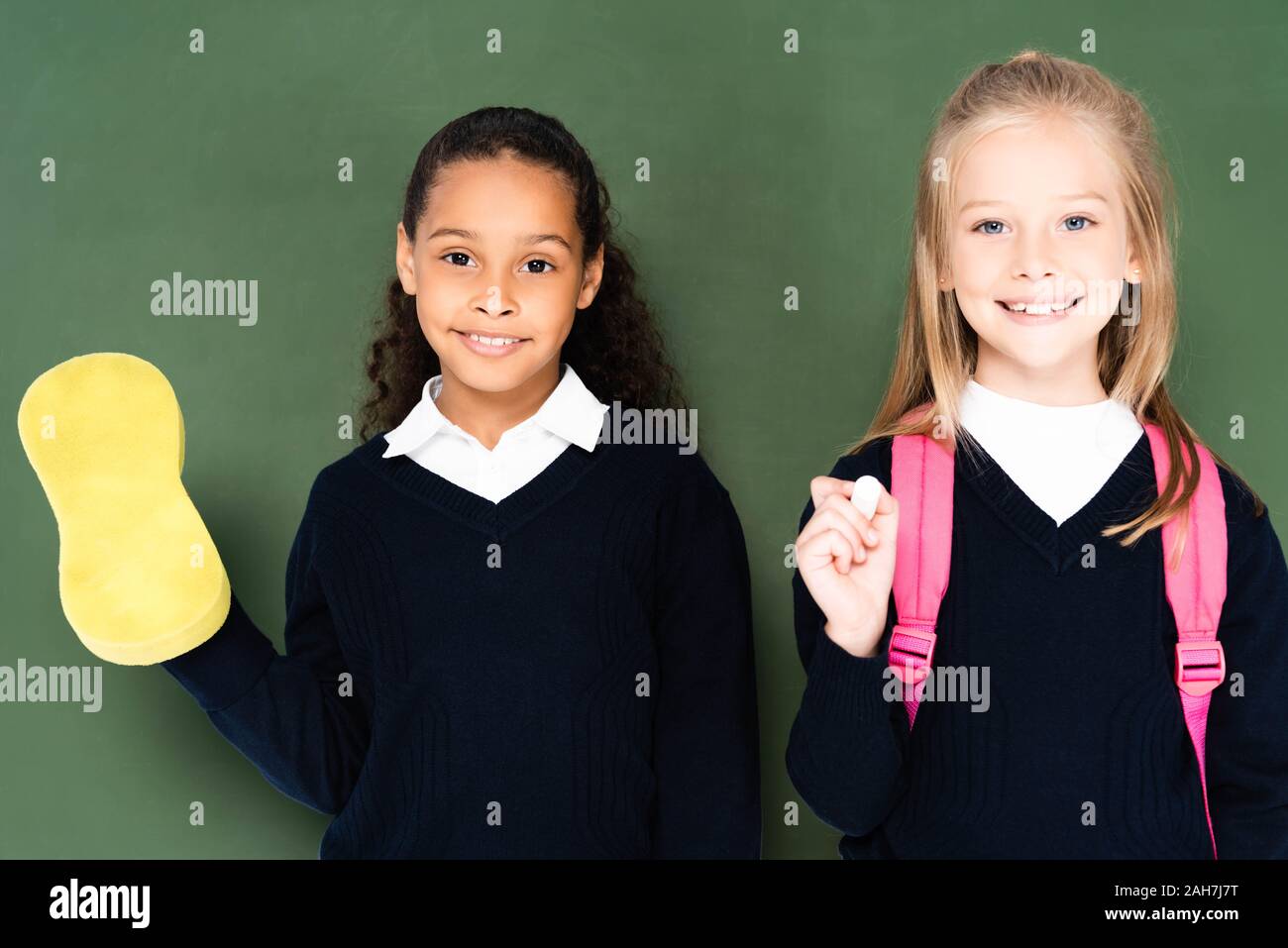 Due sorridenti studentesse multiculturale con spugna e un pezzo di gesso in piedi vicino a Lavagna Verde e sorridente in telecamera Foto Stock