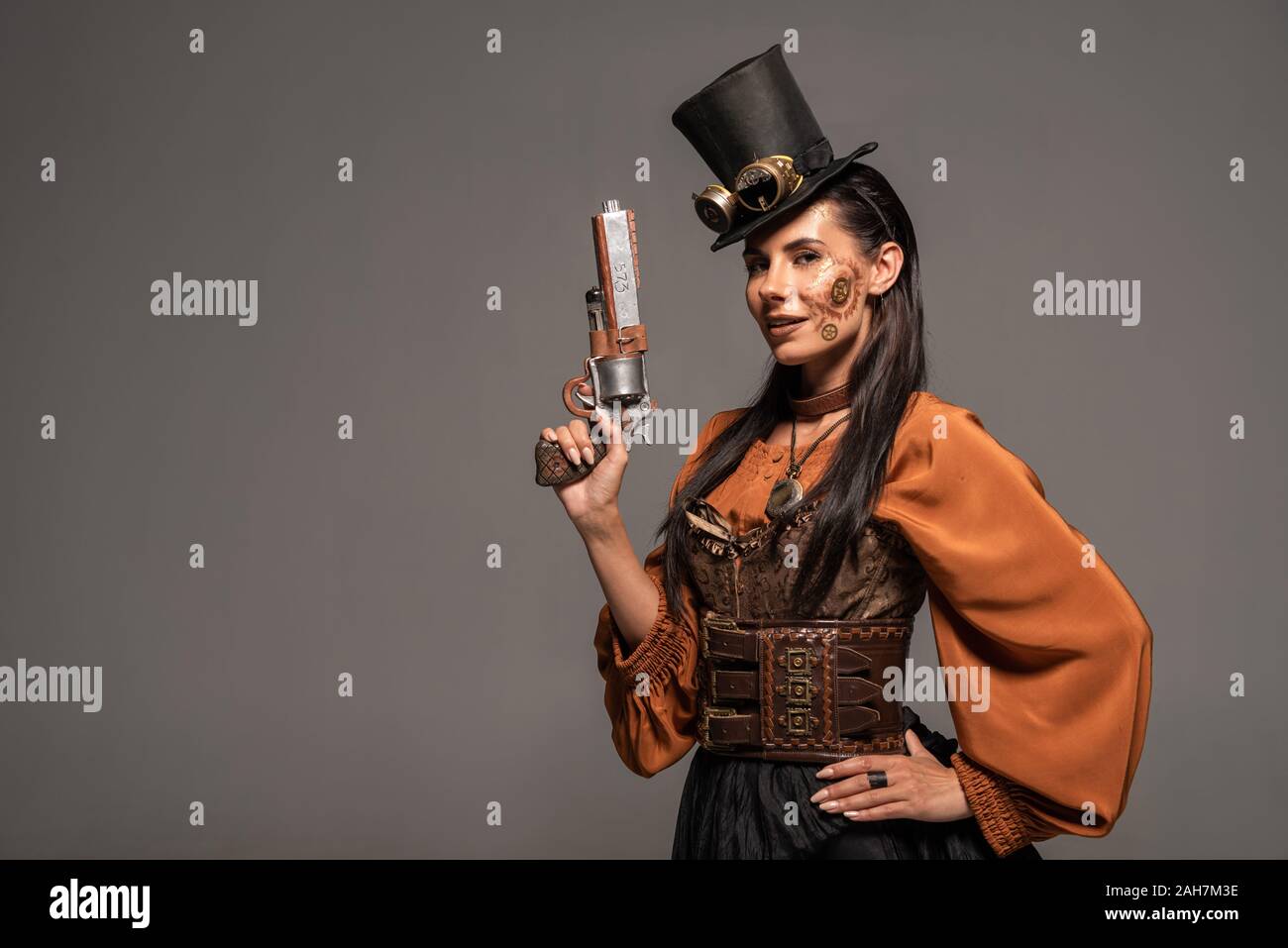 Sorridente steampunk donna in top hat con gli occhiali di protezione in piedi con la mano sul hip e pistola di contenimento isolato su grigio Foto Stock