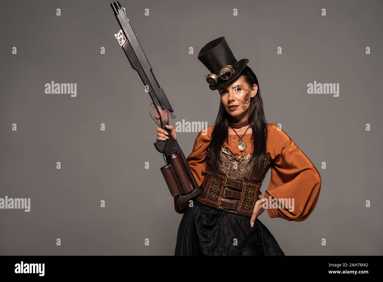 Attraente steampunk donna in top hat tenendo la pistola e in piedi con la mano sul hip isolato su grigio Foto Stock