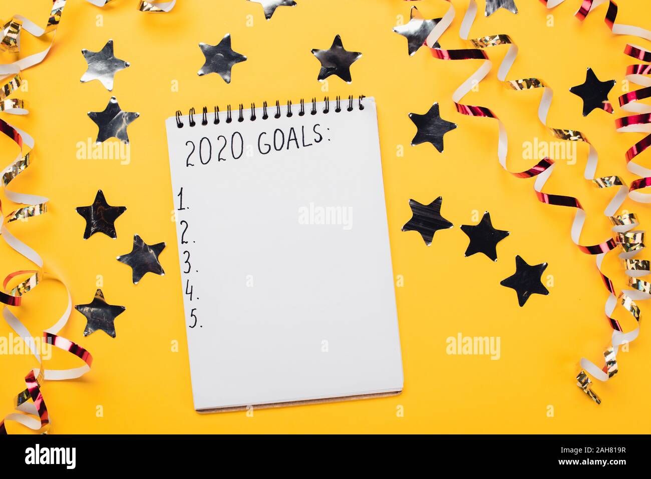 Notebook con 2020 obiettivi con vuoto di punti numerati vicino decorativo, lucenti stelle e serpentina su superficie gialla Foto Stock