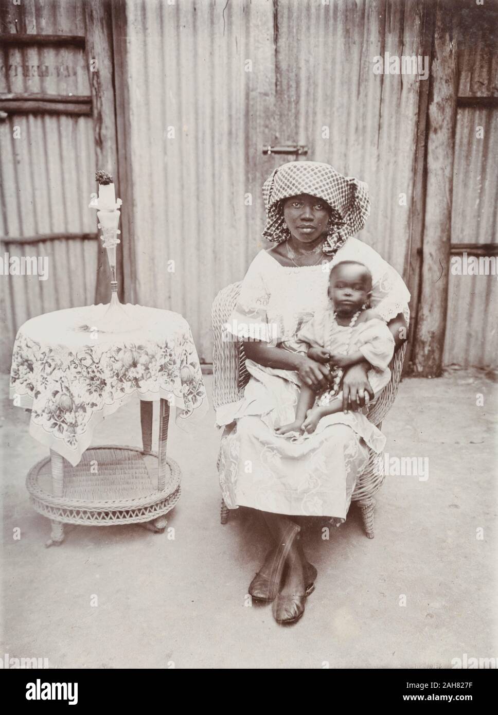 La Nigeria, la didascalia recita: "alcuni esempi del nostro cuoco della business fotografia". La donna ed il bambino sono seduto in una sedia di canna nella parte anteriore di un ferro corrugato parete e cancelli. Accanto a loro è una piccola tabella di canna con un panno floreali e vaso, [c.1920s]. 2000/098/1/115. Foto Stock