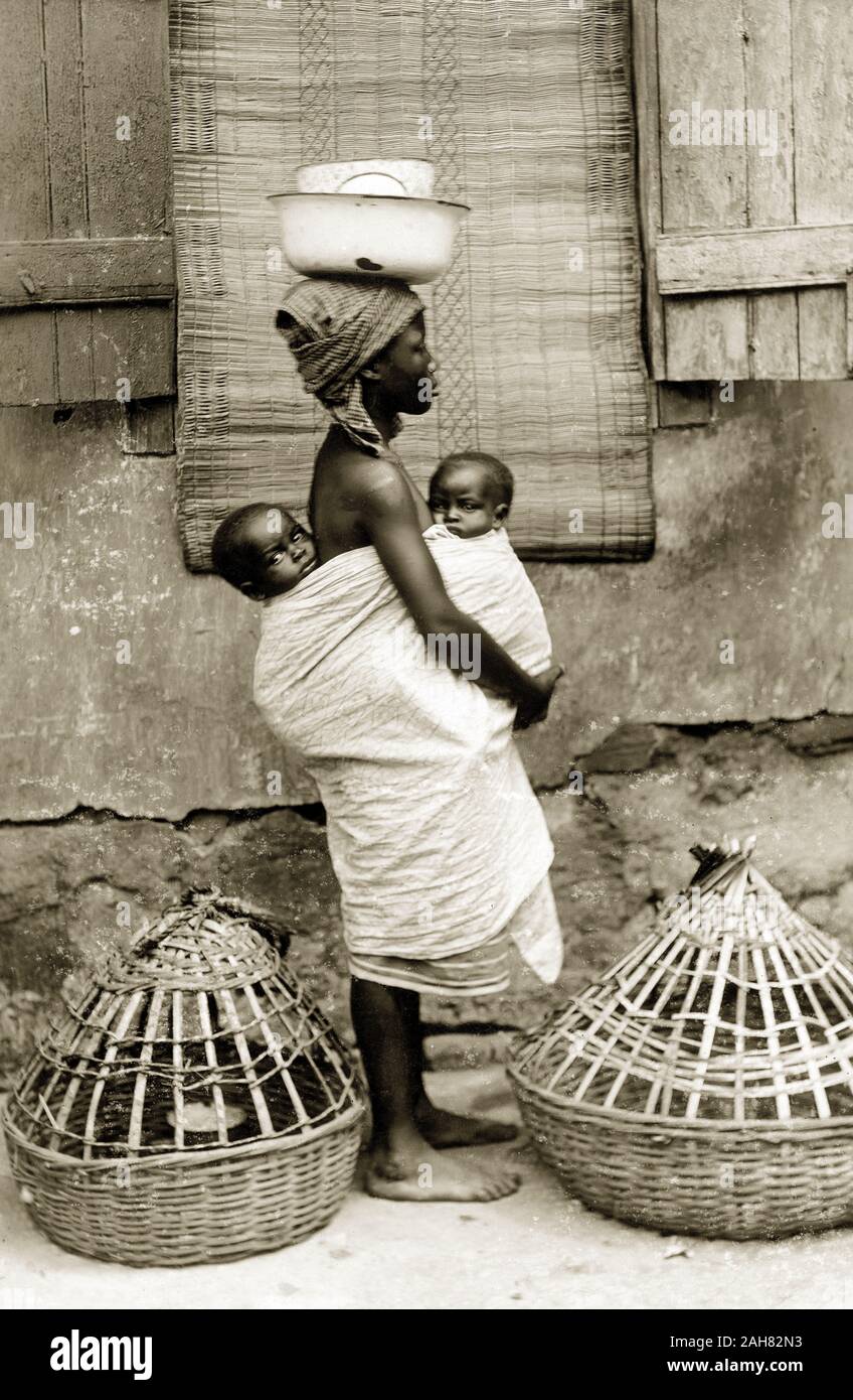 La Nigeria, la marcatura su retromarcia: "Una giovane ragazza Yoruba moglie con i suoi due 'pickins''. Precedentemente alla foto che portava un cesto all'interno dell'altro sul suo capo oltre a quello che ha sul suo capo qui. Essi sono costituiti da cestelli per polli e sono molto pesanti anche se essi non lo guardiamo. Nota il tappetino nativo appeso alla finestra utilizzata in quella casa come una cortina", [c.1920s]. 2000/098/2/82. Foto Stock