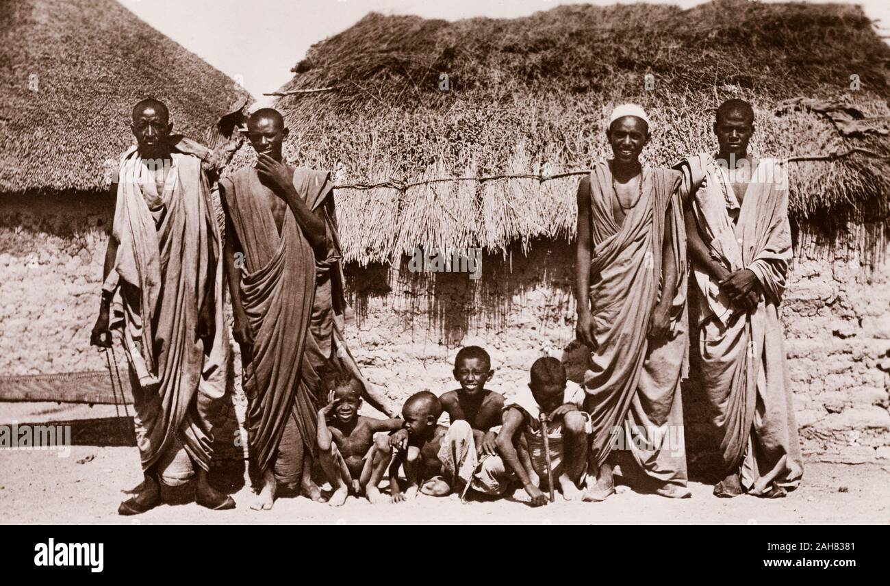Sudan, stampato didascalia recita: "L'Battaheen arabi. Pubblicato da G N Morhig, la farmacia inglese, Khartoum. Copyright 79'. Quattro uomini sudanesi pongono per la telecamera con quattro figli piccoli al di fuori di un fango-walled abitazione. Una didascalia originale li identifica come "Battaheen [Bataheen] gli arabi", uno dei più distinti gruppi tribali residenti in stati del nord del Sudan, [c.1906]. 2003/222/1/2/48. Foto Stock