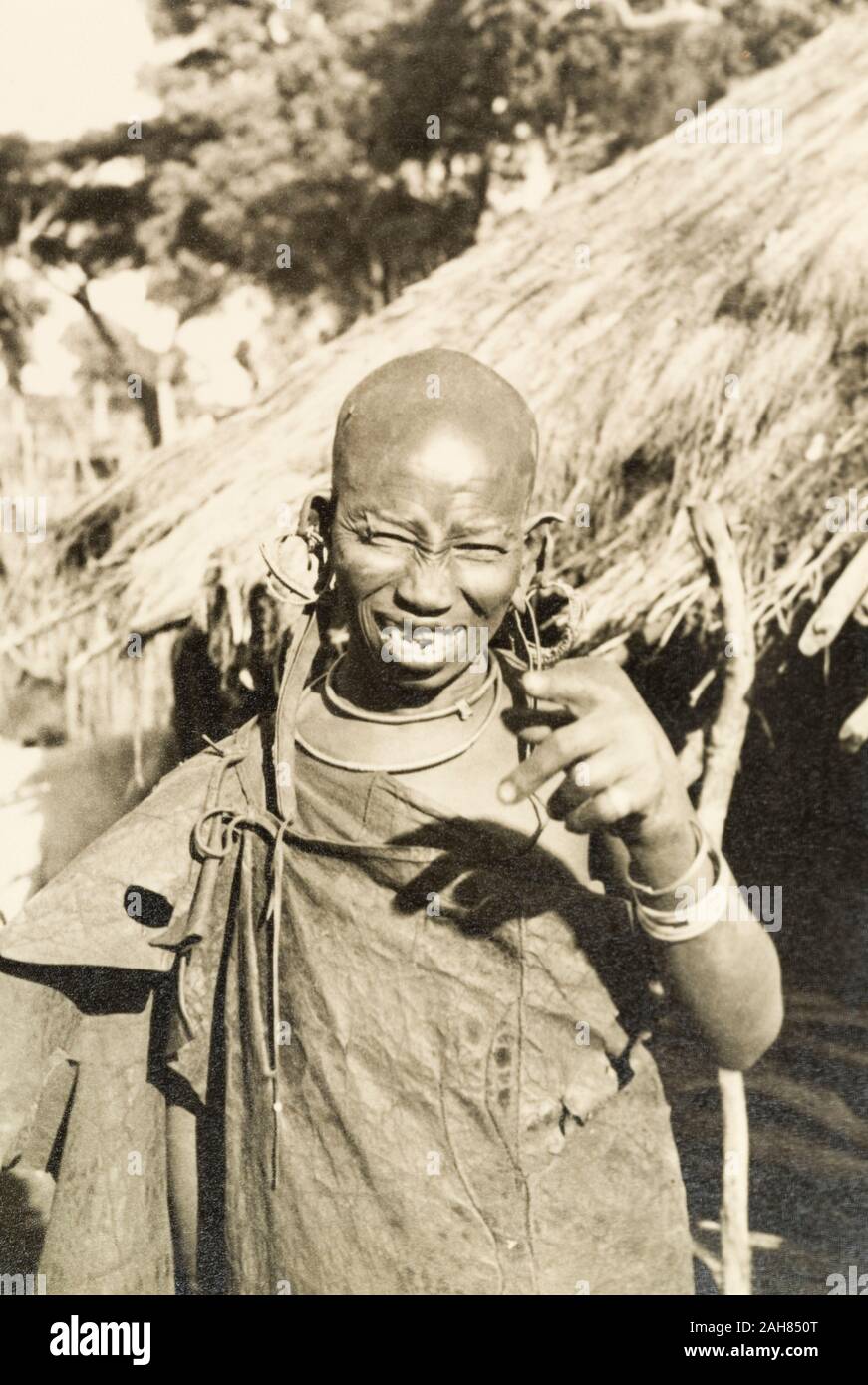 Kenya, Ritratto di una sorridente donna Kikuyu gesticolando alla fotocamera. La sua testa è rasato, che indica il suo stato come una donna sposata, e indossa un numero di orecchini a cerchio. Manoscritto originale didascalia: Una vecchia donna Kikuyu.Circa 1934, circa 1934. 1995/076/1/2/5/37. Foto Stock