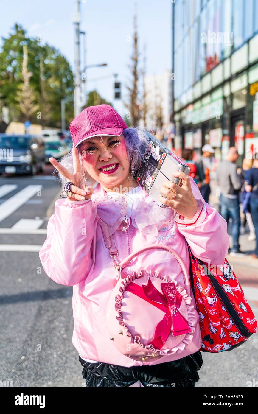 Kawaii Deocra-Chan Japanese street fashion. Donna matura vestita in rosa con Hello Kitty borsa. Sorridente e dando 2 dito gesto di pace. Harajuku. Foto Stock