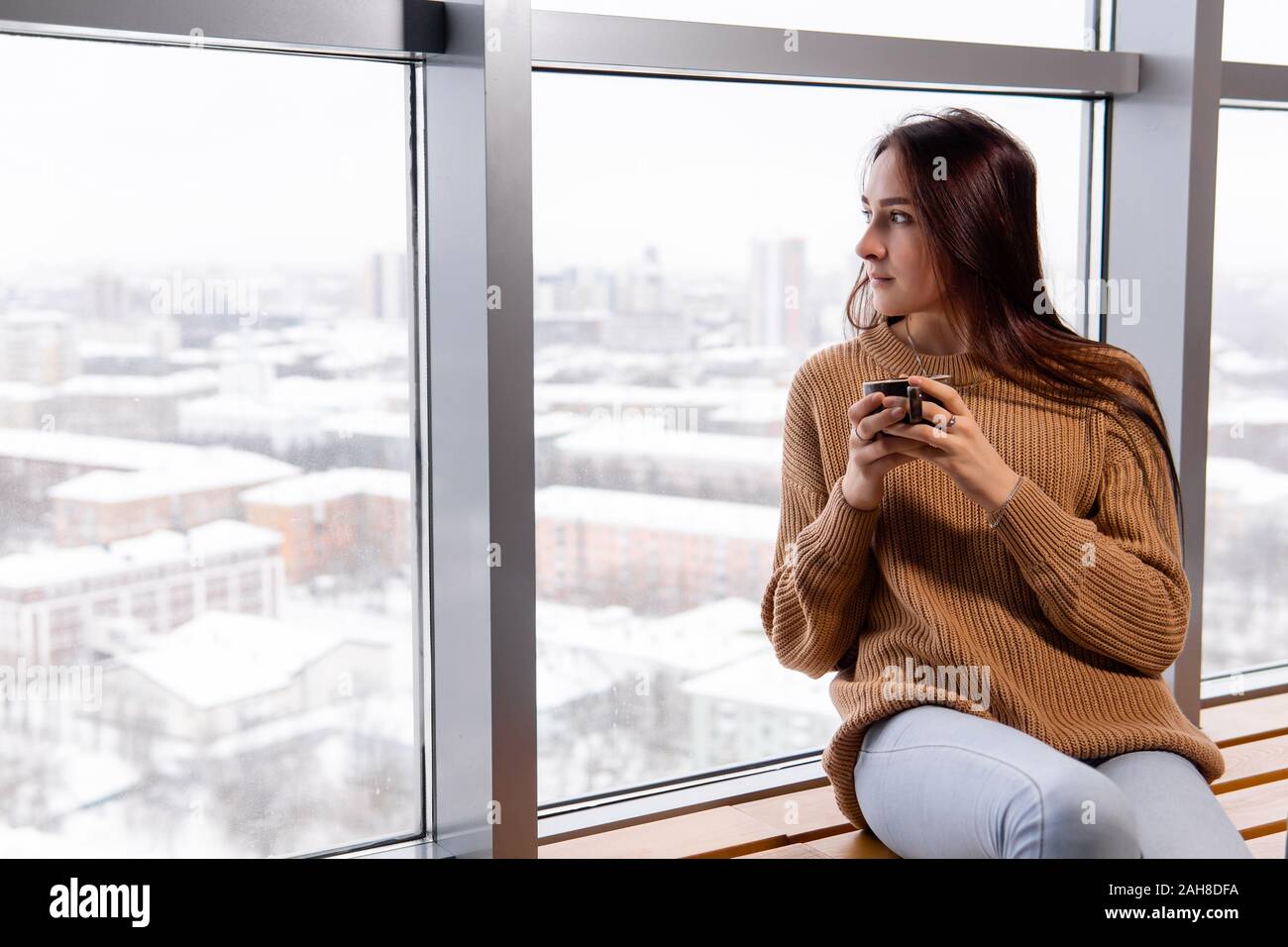 Giovane donna con una tazza di caffè si siede dalla finestra al piano attico di un edificio alto, guardando verso il basso sulla città in una giornata invernale Foto Stock