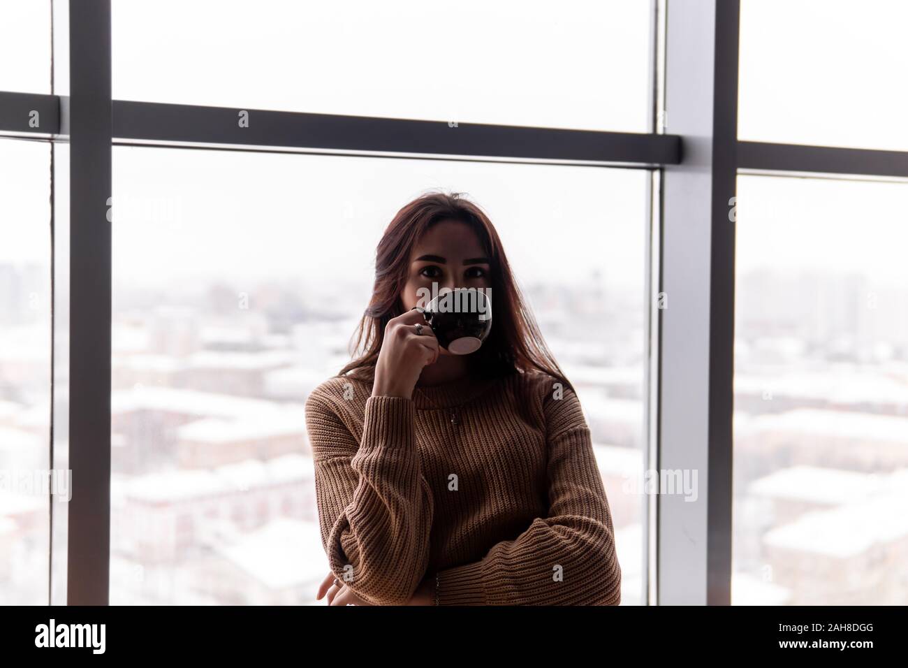 Giovane donna bere il caffè seduta dalla finestra al piano attico di un edificio alto, contro la città su di una giornata invernale Foto Stock