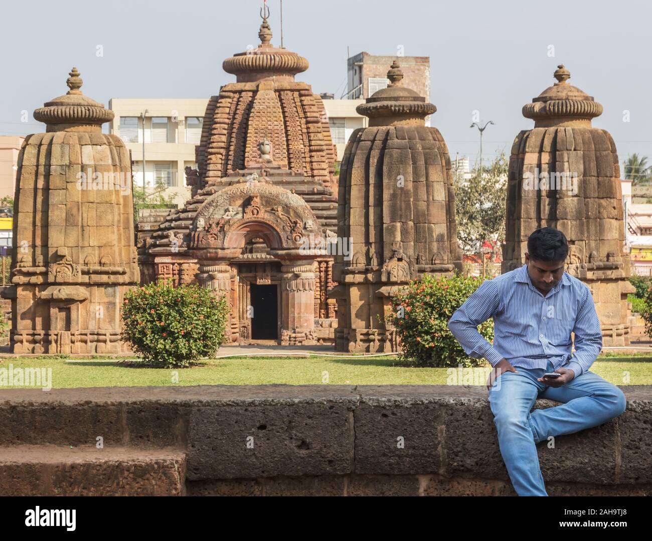 Bhubaneshwar, Orissa, India - febbraio 2018: Un uomo siede su un muro di pietra e guarda al suo telefono con gli antichi templi di Mukteshwar sullo sfondo. Foto Stock