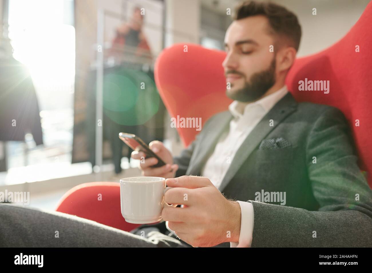 La mano di giovani ben vestito uomo d affari con tazza di tè rilassanti in poltrona rossa Foto Stock
