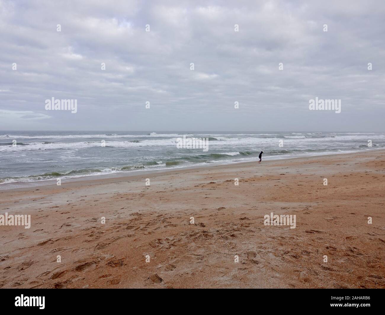 Giovane in felpa con cappuccio altrimenti sulla spiaggia vuota su un blustery giorno di dicembre con cappucci bianchi e le onde, Saint Augustine Beach, Florida, Stati Uniti d'America. Foto Stock