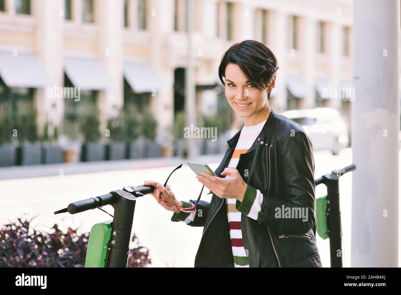 Cavalletti femmina da scooter elettrico nella città e guarda la fotocamera a sorridere - telefono cellulare in mano per utilizzare app in affitto - ore diurne Foto Stock