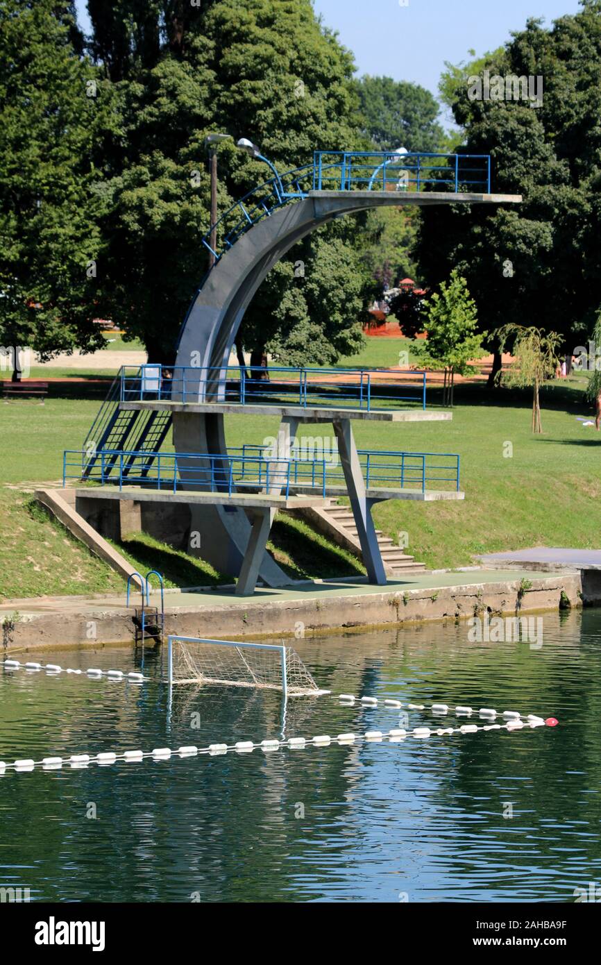 Tre livelli di altezza vecchio cemento trampolino piattaforma con blu di recinzione metallica a livello locale in riva al fiume circondato da erba e alberi in background Foto Stock