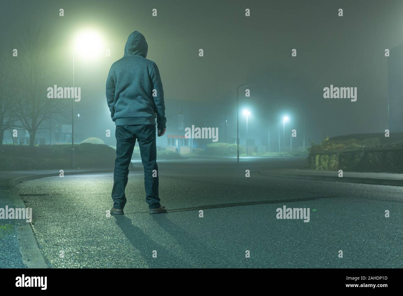 Una misteriosa figura con cappuccio con torna alla telecamera, in piedi su una strada in una luce industrial area urbana. Su un moody, foogy, inverni di notte Foto Stock