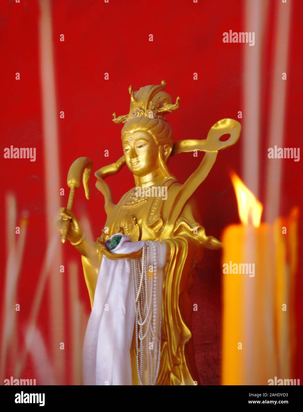 Statua dorata di una dea buddista contro la parete rossa. Le candele e i bastoncini di incenso in primo piano. Foto Stock