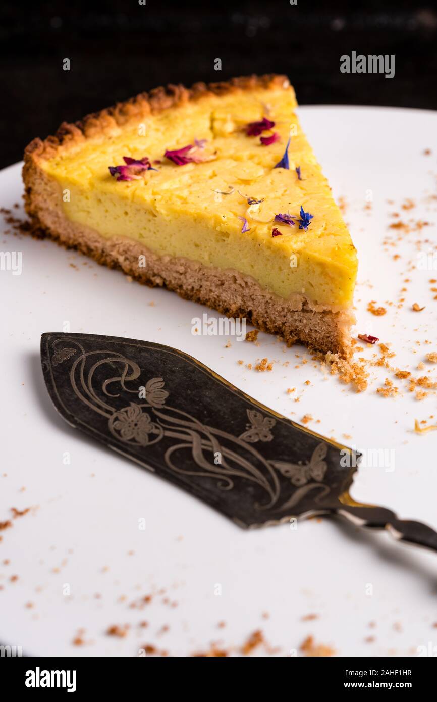 Un pezzo di vegan Cheesecake al limone, guarnito con petali di fiori commestibili, su una piastra bianca ornato di torta server e alcune briciole - close-up, verticale Foto Stock