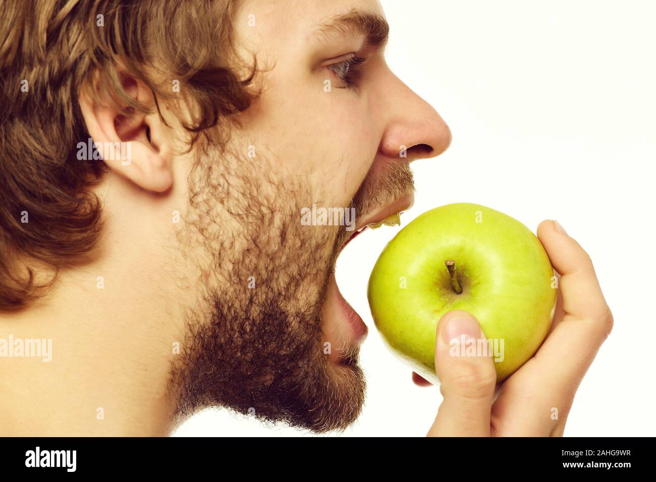 Ragazzo con barba corta cercando di mordere grande mela verde sta guardando con la bocca aperta, in vista laterale Isolato su sfondo bianco. Cibo sano modello Foto Stock