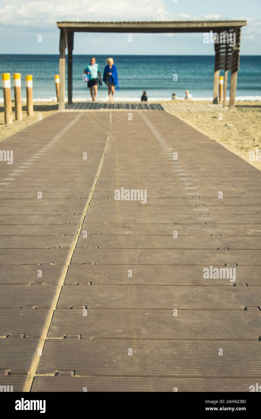 Coppia offuscata sullo sfondo su una passeggiata in spiaggia di legno a Playa del Ingles, Gran Canaria. I turisti godono di vacanza sulla spiaggia in Isole Canarie, Spagna Foto Stock