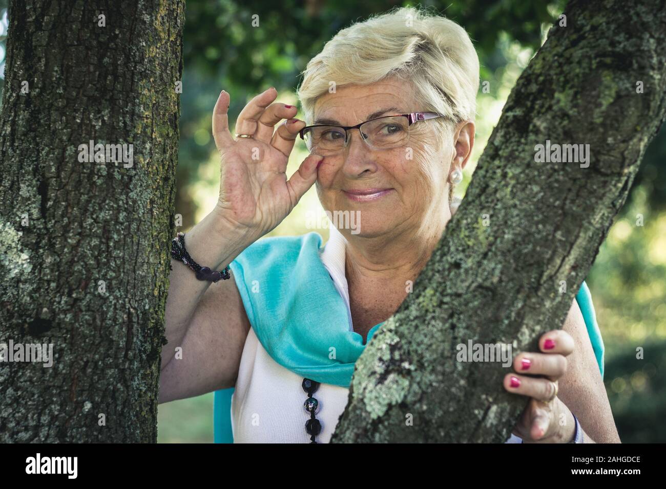 Blonde elder donna pone tra i tronchi di alberi nel parco. Senior Signora sorride tenendo occhiali e guardando la fotocamera Foto Stock