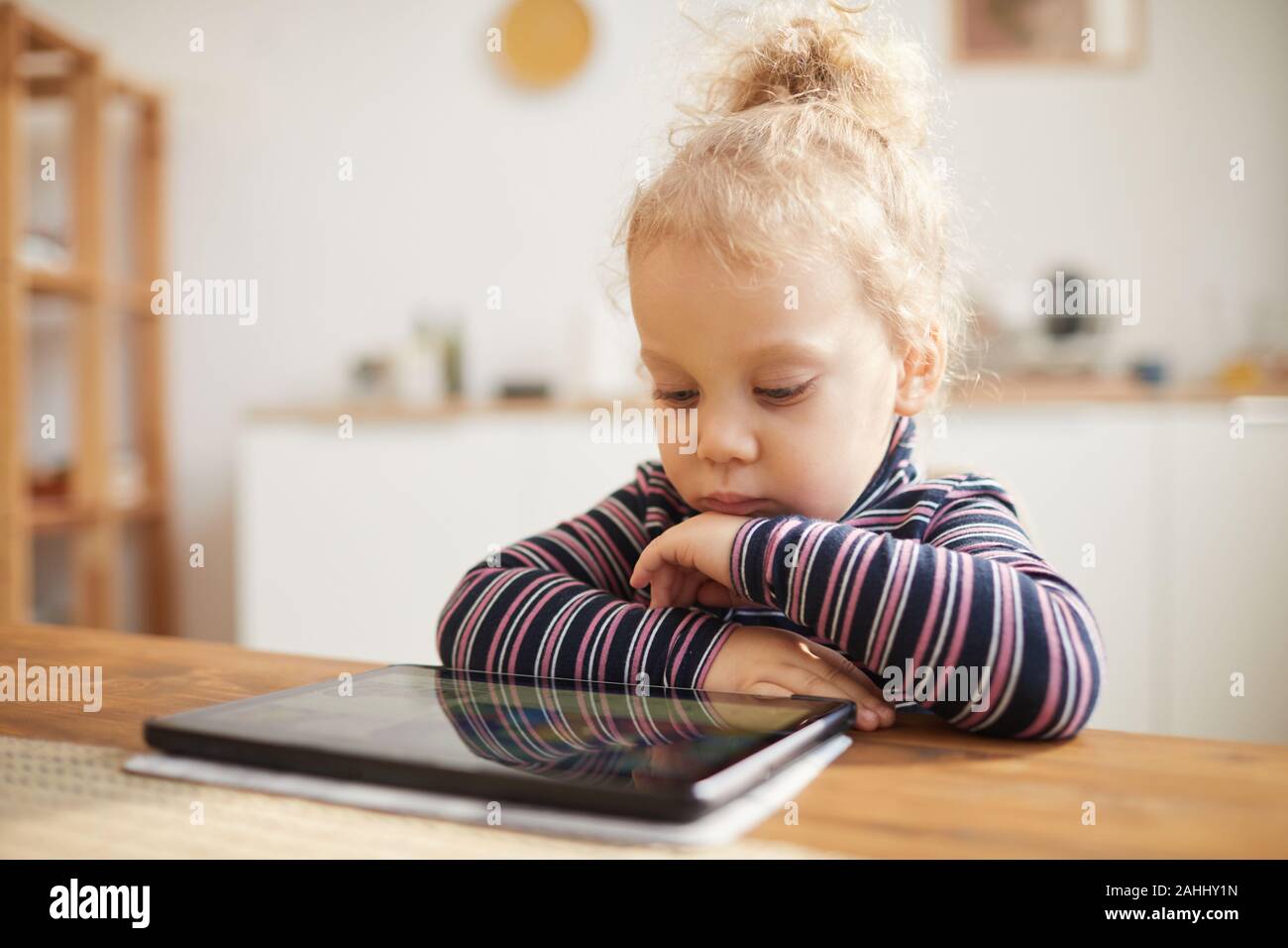 In tonalità calde ritratto di carino bambina guardando a tavoletta digitale mentre è seduto al grande tavolo in accogliente cucina, spazio di copia Foto Stock