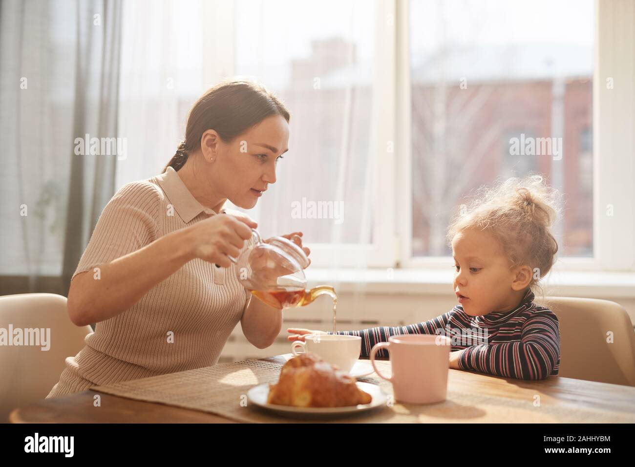 Vista laterale verticale della moderna madre versando il tè nero per cute bambina mentre gustate la prima colazione a casa seduti contro la finestra nella luce solare Foto Stock