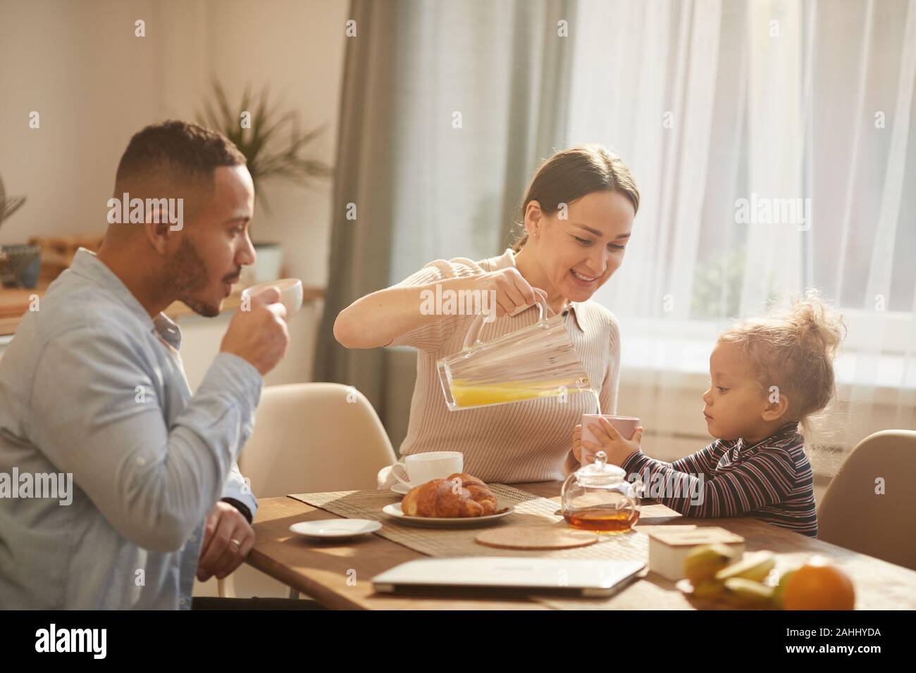 In tonalità calde ritratto della moderna famiglia felice gustando la prima colazione a casa seduti al tavolo da cucina in presenza di luce solare Foto Stock