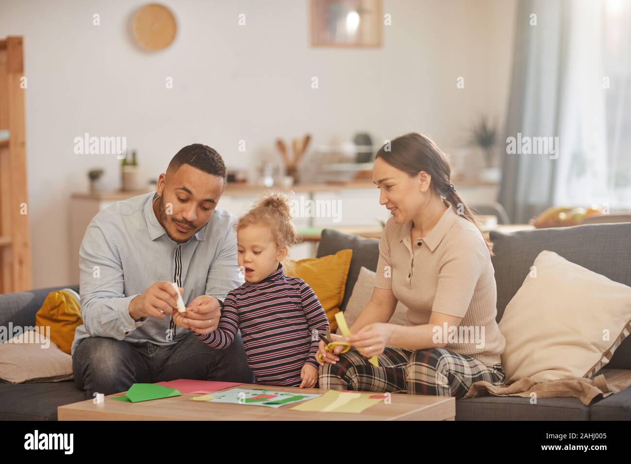 Dai toni caldi ritratto di felice mixed-gara famiglia giocando con incantevole piccola figlia in casa accogliente interno Foto Stock