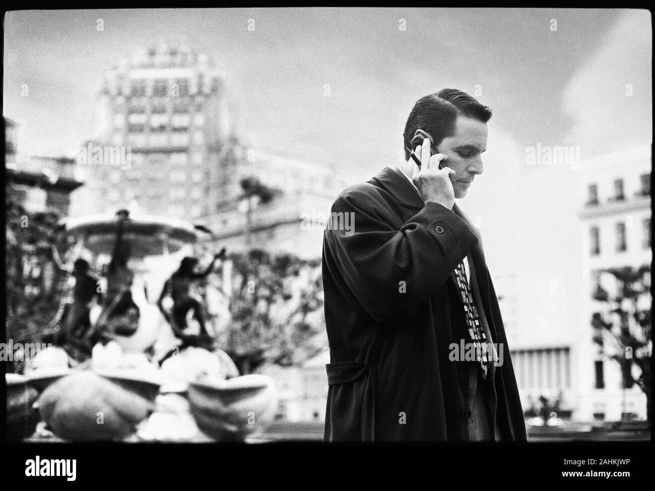 Vista laterale di un uomo che parla sul suo telefono cellulare con una fontana in background. Foto Stock