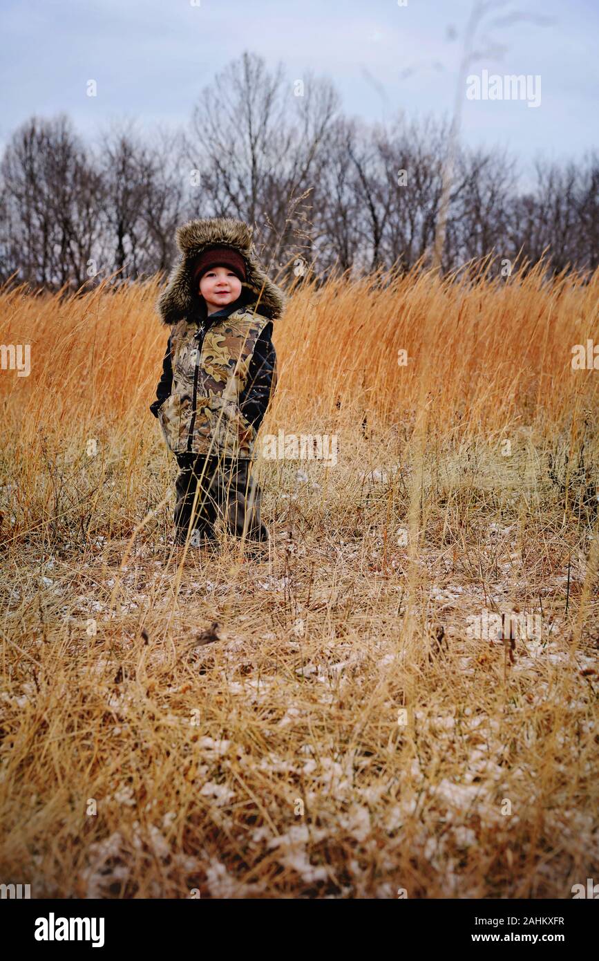 Little Boy indossando giubbotto camoflauge e foderata in pelliccia di trapper hat in un campo Paese Foto Stock