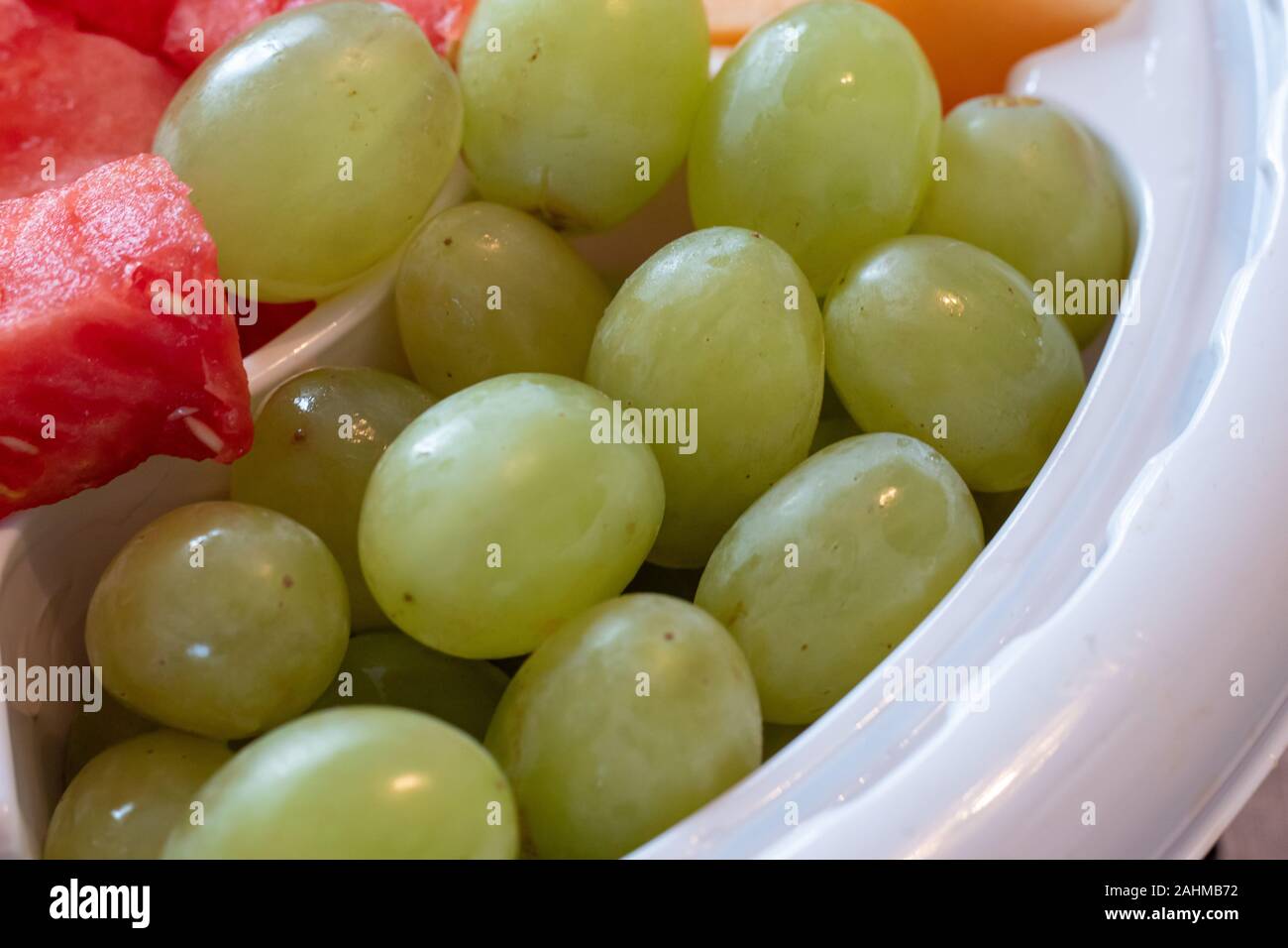 Uva verde sono serviti in un bianco di plastica del vassoio di frutta e sono visti da vicino. Anguria e le fette di melone può essere visto in corrispondenza dei bordi. Foto Stock