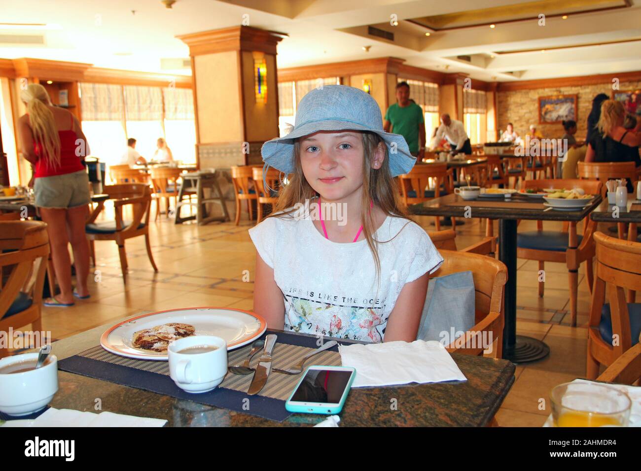 Bambino moderno sorridere mentre sedendo nel ristorante. Bambina in hat è a pranzo in ristorante. La ragazza di hat in posa mentre mangia Foto Stock