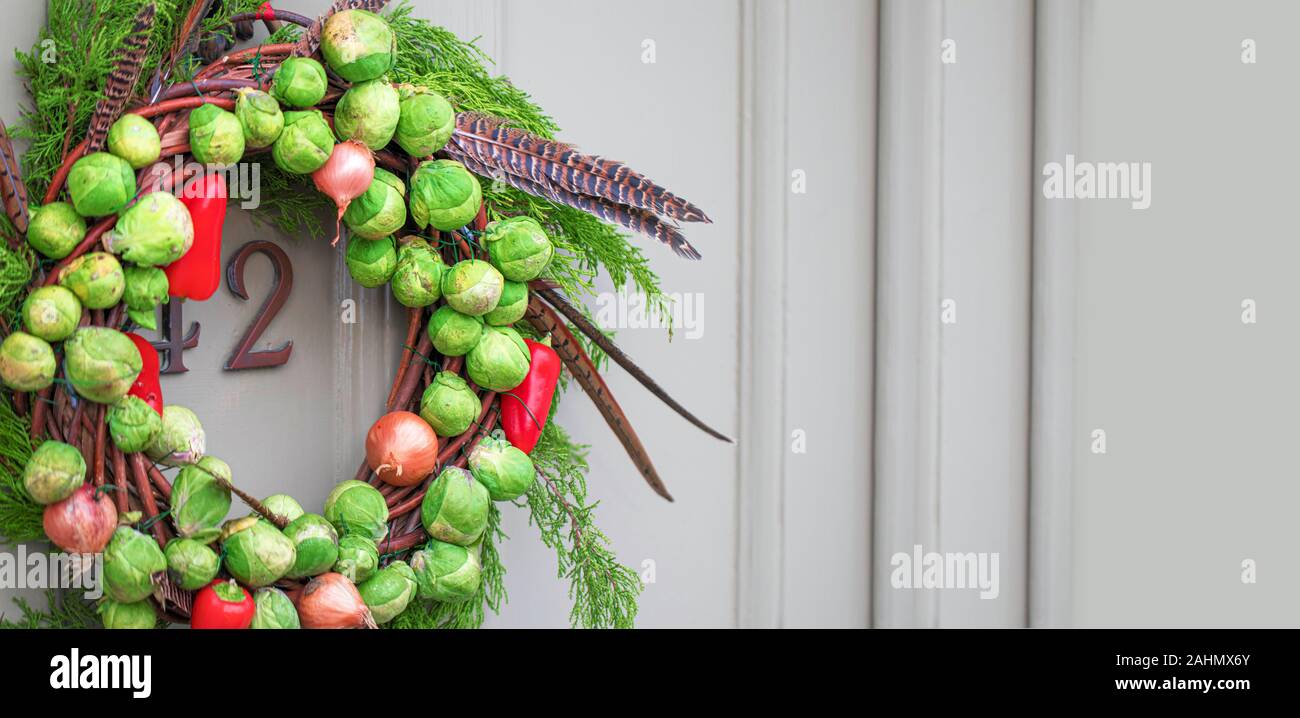 Decorateive festosa vegetali fatti a mano ghirlanda fatta di i cavoletti di Bruxelles, cipolle, Chilis, foglie sempreverdi e Piume fagiano appeso su una porta vecchia Foto Stock