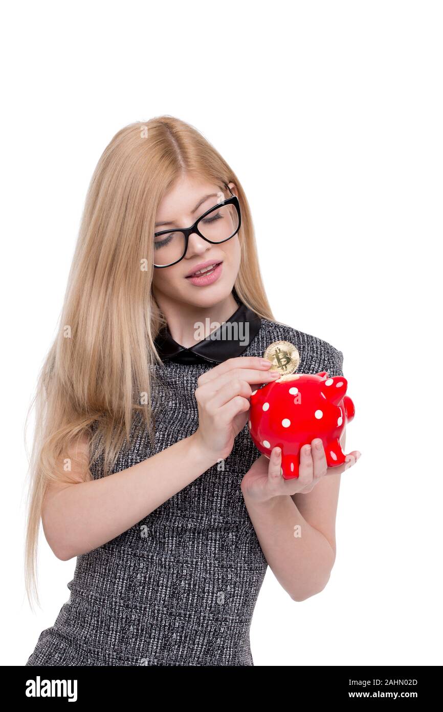 Bionda e giovane imprenditrice goccia bitcoin in punteggiata rossa salvadanaio, isolato su bianco Foto Stock