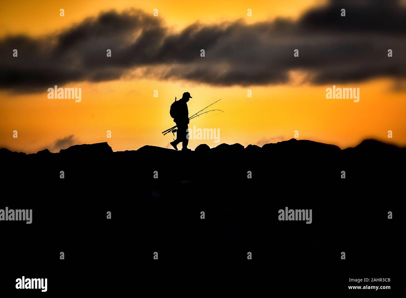 Silhouette di uomo con poli di pesca a piedi attraversata rocce del molo al intensa arancione tramonto Foto Stock