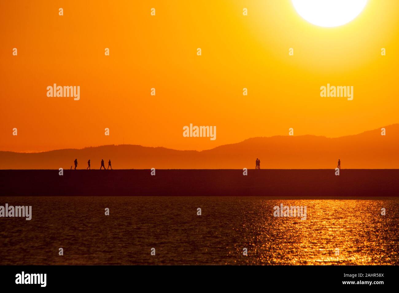 Stagliano walkers su un molo al momento di un tramonto dorato, con il mare in primo piano e le montagne sullo sfondo Foto Stock