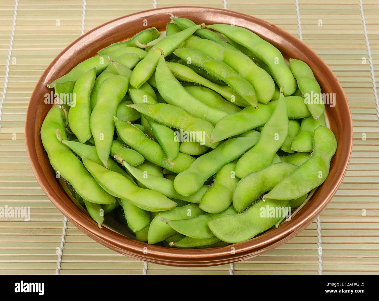 Edamame bolliti in una ciotola marrone su un tappetino di bambù. Fagioli di soia verde, anche maodou, cotto in acqua salata. Un piatto di lato e gustosi snack. I fagioli di soia, Glycine max. Foto Stock